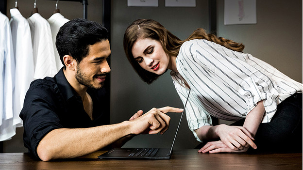 Man holds index finger on laptop screen and woman peeks around the corner.