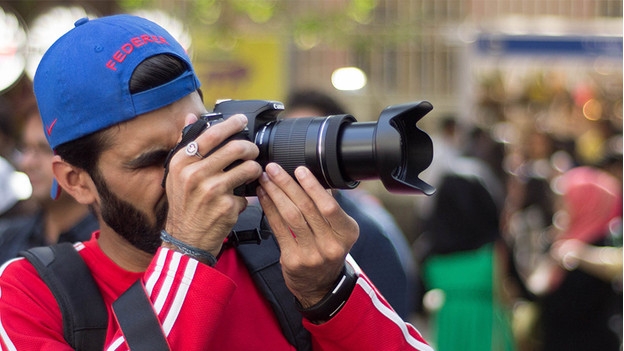 Man met rode trui en blauwe pet maakt foto met camera. 