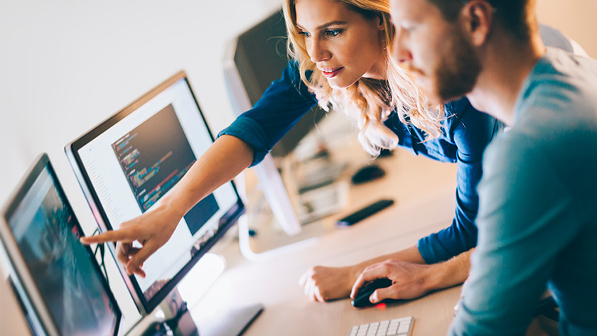 Vrouw wijst naar monitor computer op bureau. Man kijkt met computer muis in hand toe. 