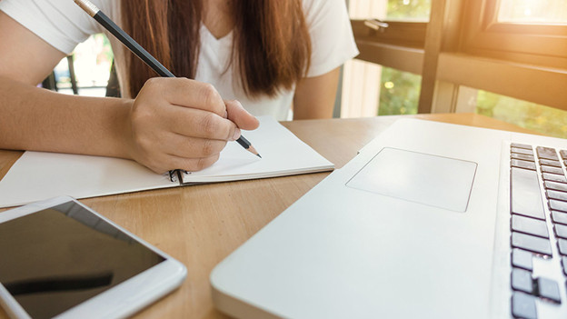 Woman writes in notebook while she works on a laptop behind a desk.